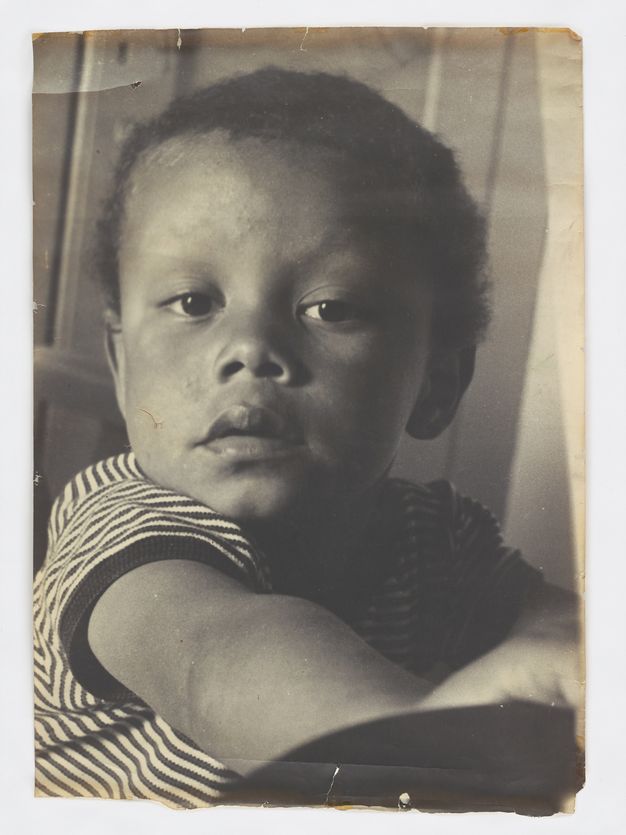 Black and white portrait of a young child with short hair, gazing directly at the camera. The child is wearing a striped shirt and appears to be in a thoughtful or contemplative mood. The photograph exhibits signs of aging, with visible creases and slight discoloration on the edges, suggesting it is an older print. The lighting is natural, highlighting the child's facial features and expression.