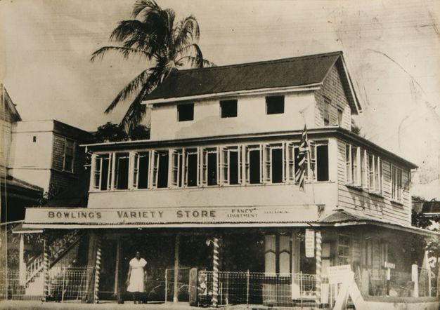 Black and white photograph depicting a two-story colonial-style building labeled "Bowling's Variety Store." The structure features wooden siding, large windows, and a gabled roof. A staircase is visible on the left. A person in a white dress stands in front of the building, which is surrounded by a low wire fence. Palm trees are visible in the background, suggesting a tropical setting. The image captures early 20th-century architectural and cultural elements.